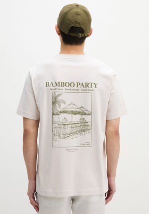 Mężczyzna noszący białą koszulkę z zielonym napisem "Bamboo Party" i ilustracją bambusowego baru na plecach, w zestawieniu z oliwkową czapką.