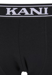 Melni vīriešu bokseršorti ar plašu jostu, uz kuras ir izteikts balts "KANI" teksts un baltas svītras uz melna fona.