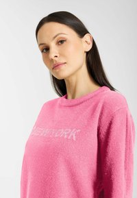 Różowy sweter z miękkiej, teksturowanej tkaniny z napisem "NEW YORK" w błyszczących literach na klatce piersiowej. Okrągły dekolt i luźny krój.