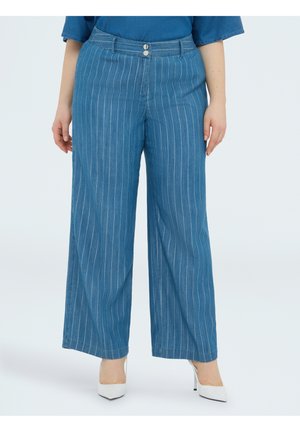 Frau trägt eine hoch taillierte, blaue Nadelstreifen-Hose mit weitem Bein und weiße spitze High Heels, steht vor einem einfarbigen Hintergrund.