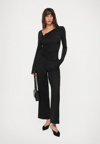 Fekete ráncolt aszimmetrikus felsőt, széles lábú nadrágot, fekete szandált viselő nő, aki egy fekete táskával áll egy sima fehér háttér előtt.