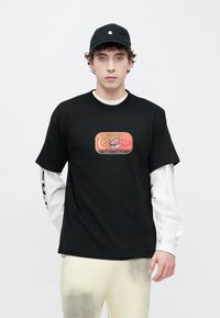 Camiseta negra de algodón con un logo colorido impreso en el frente, sobre una camisa blanca de manga larga; combinada con pantalones de color claro.