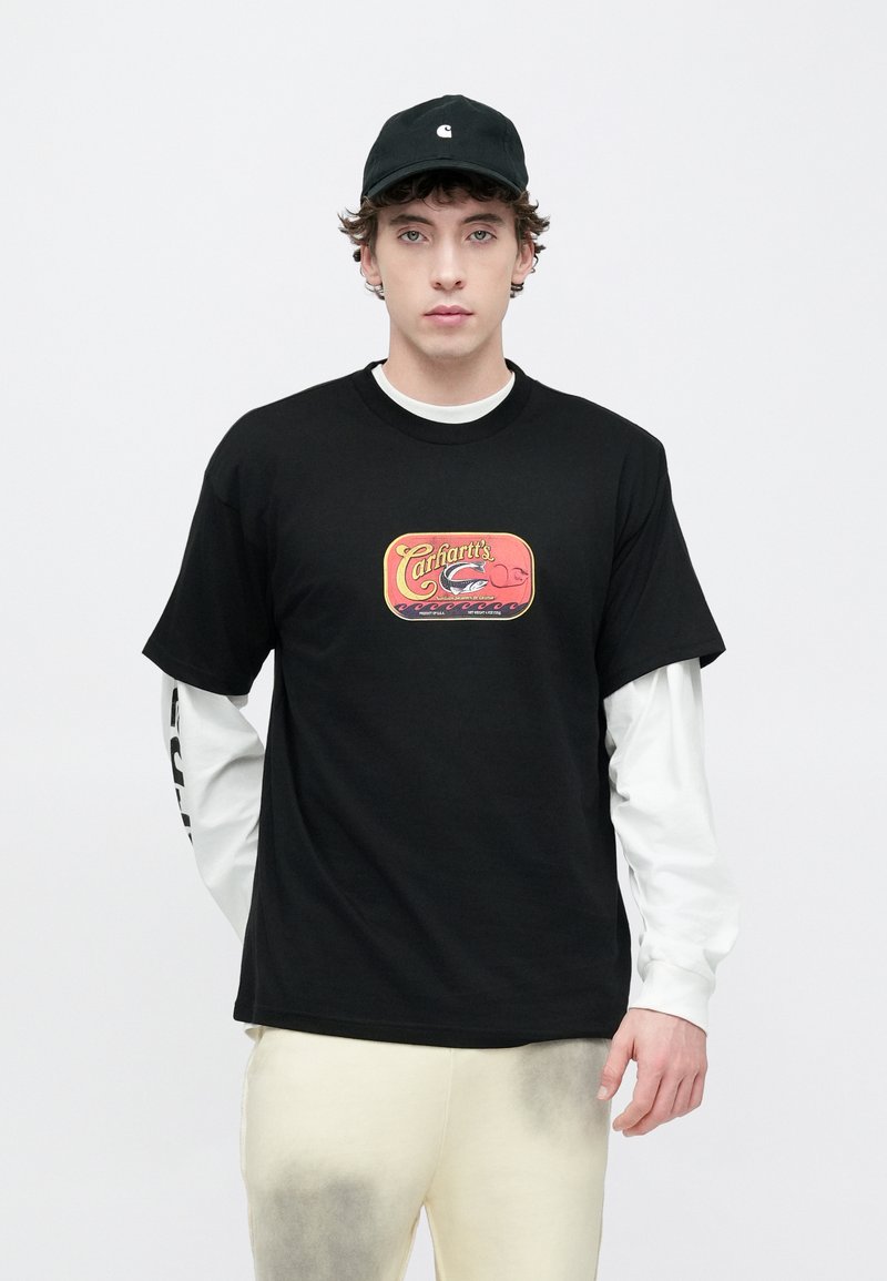 Zwart katoenen T-shirt met een kleurrijk bedrukt logo op de voorkant, gedragen over een wit shirt met lange mouwen; gecombineerd met lichtgekleurde broek.