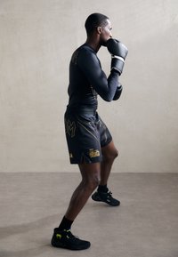 Mann in schwarzer Sportbekleidung und Boxhandschuhen, der in einer Kampfposition auf einem grauen Boden und vor einer schlichten Wand steht.