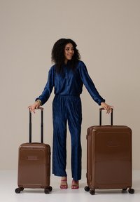 Two hard-shell suitcases in brown with vertical ribbed design, held by a person in a dark blue outfit, standing on a light background.