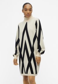Crème trui jurk met zwart zigzagpatroon, hoge opstaande kraag en geribbelde manchetten. Zachte textuur, relaxte pasvorm, lengte tot halverwege de dij.