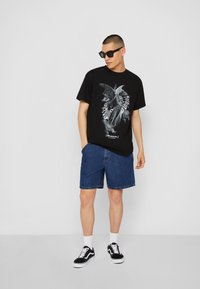 Svart grafisk t-skjorte med et engeldesign, paret med blå denimshorts. Brukt med svarte joggesko og solbriller. Enkel bakgrunn.