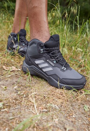 Nogii w szarych i czarnych butach trekkingowych Adidas Terrex stoją na ziemnej ścieżce otoczonej trawą i roślinnością.