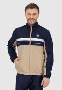 Giacca navy e khaki con zip completa, colletto alto e strisce bianche in evidenza sul petto. Presenta polsini elasticizzati e tasche laterali.