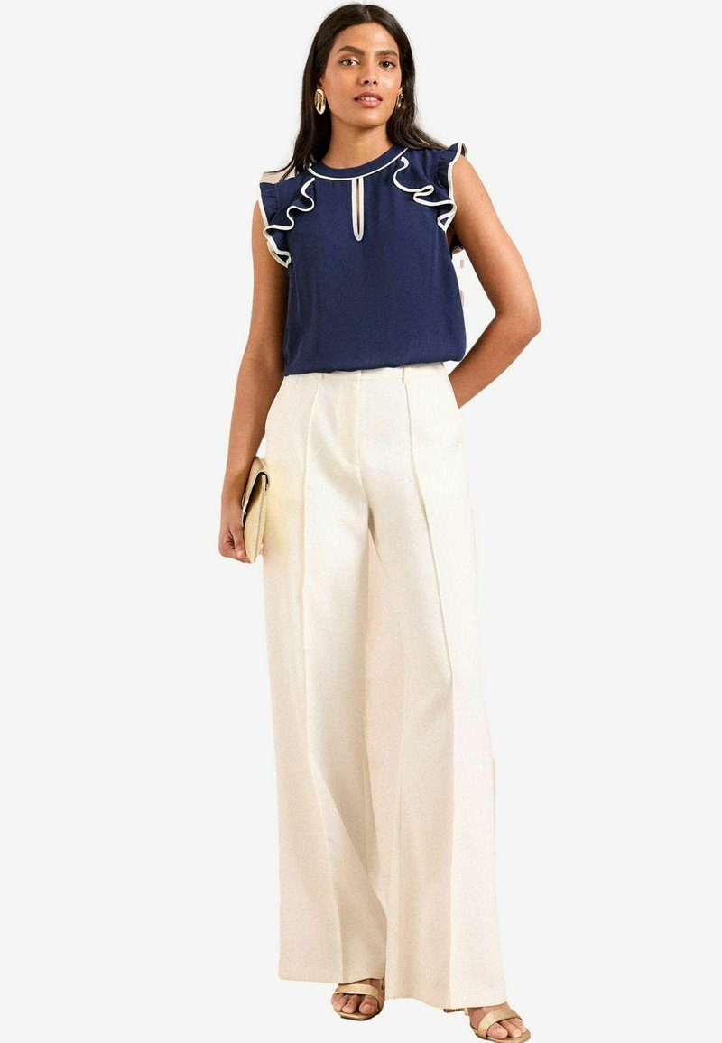 Blusa blu con maniche arricciate e bordo bianco, abbinata a pantaloni larghi color crema. Gli accessori includono una piccola pochette e sandali con cinturino.