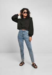 Femme aux cheveux bouclés portant un sweat-shirt noir court, un jean taille haute bleu, des sandales noires et des lunettes de soleil sombres, se tenant avec une main sur la tête.