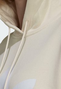 Krémová mikina s kapucí z měkkého materiálu, vybavená stahovací šňůrkou na kapuci a velkým bílým logem na přední straně. Pohled zblízka zdůrazňuje detaily tkaniny.