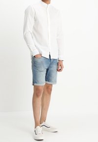 Camicia bianca in lino a maniche lunghe con colletto alla francese, abbinata a pantaloni corto in jeans arrotolati. Sneakers bianche con dettagli blu. Outfit casual.