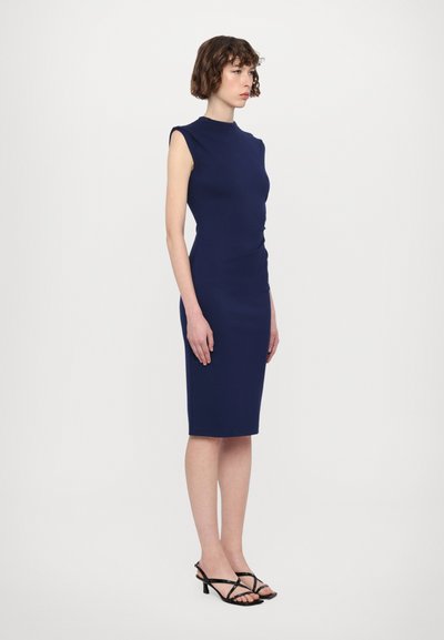 Rochie ajustată de culoare bleu marin, cu design fără mâneci, decolteu înalt și detalii plisate la talie; purtată cu sandale cu toc negre.