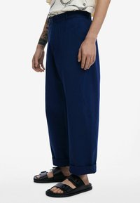 Pantaloni ampi blu navy realizzati in tessuto liscio, caratterizzati da un design tapered, tasche laterali e orli a risvolto, abbinati a sandali neri.