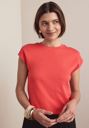 Donna con capelli castani corti che indossa una maglietta corallo a maniche a cappuccio e orecchini d'oro, che sorride leggermente e guarda di lato, con le mani incrociate davanti.