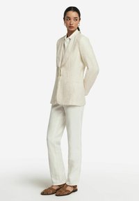 Krämfärgad blazer med textur och skräddarsydd passform, i kombination med ljusa byxor och bruna remsandaler med en minimalistisk design.