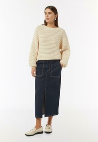 Beżowy sweter z dzianiny z wzorem teksturowym, w zestawieniu z ciemnoniebieską spódnicą dżinsową z rozcięciem z przodu i kontrastowym przeszyciem. Białe mokasyny.