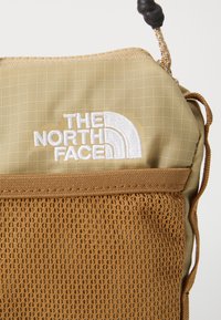 Tan stoffpose med nettbunn, med en hvit brodert logo. Drawstring-lukking med en teksturert svart perle for stramming.