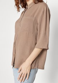 Donna che indossa una camicia beige con bottoni e tasca sul petto e jeans azzurri chiari, visibile dalle spalle ai fianchi su uno sfondo bianco.