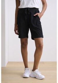 Černé bavlněné šortky s tkaničkou v pase, bočními kapsami a ohrnutými lemy, spárované s bílými sportovními botami na dřevěné podlaze.