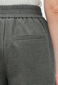 Detailný záber osoby v tmavosivých elastických nohaviciach s elastickým pásom a zadnou vreckom, zobrazenie časti ruky a bielej košele.