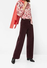 Pantalon long en velours côtelé bordeaux avec poches latérales, assorti à une chemise à motifs rose à boutons et un pull texturé rose noué autour des épaules.