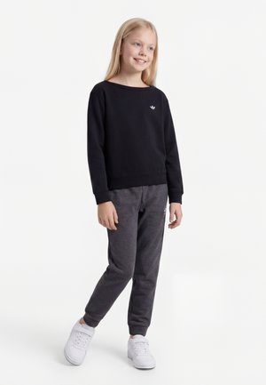 Smilende pige med langt blondt hår iført en sort langærmet trøje, mørkegrå joggingbukser og hvide sneakers, stående på en hvid baggrund.