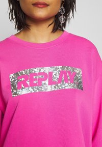 Sudadera rosa con un gráfico plateado y brillante de "REPLAY" en la parte frontal. Tiene un escote redondeado y un ajuste holgado.