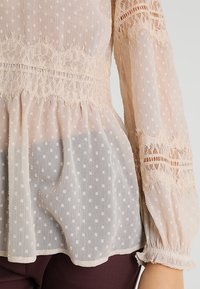 Transparente cremefarbene Bluse mit Punkten, Spitzendetails an den Ärmeln und der Taille, langen Ärmeln und einem gerafften Saum. Leichter Stoff.