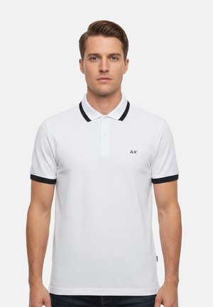 Uomo che indossa una polo bianca con rifiniture nere su colletto e maniche, in piedi davanti a uno sfondo chiaro uniforme.