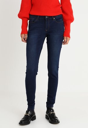 Personne portant un jean skinny bleu foncé, des mocassins en cuir noir avec des détails à franges, et un pull côtelé rouge avec des manches ballon.