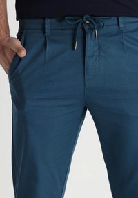 Chino teal en tissu texturé, avec une taille à cordon de serrage, deux poches latérales et une coupe ajustée, présentant une finition lisse.
