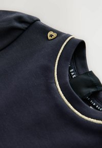 Donkerblauwe shirt met korte mouwen, voorzien van een gouden hartvormige knoop en een metalen rand langs de halslijn. Zachte katoenstofstructuur.