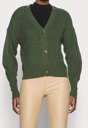 Persona que lleva un cárdigan de punto acanalado verde oscuro con tres botones sobre una camiseta negra y pantalones beige ajustados.