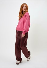 Růžová oversize mikina s kulatým výstřihem, kombinovaná s lesklými bordó kalhotami s širokými nohavicemi. Model má na sobě světle zbarvené tenisky.