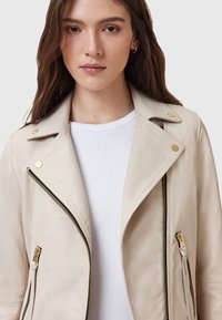 Giacca da moto in pelle beige con zip frontale e tasche laterali, hardware dorato e colletto a risvolto. Indossata sopra una camicia bianca.