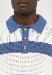 Pletená polo košile v bílé barvě s modrými vodorovnými pruhy. Obsahuje modrý žebrovaný límec a tři kulaté knoflíky u krku. Texturovaná tkanina.