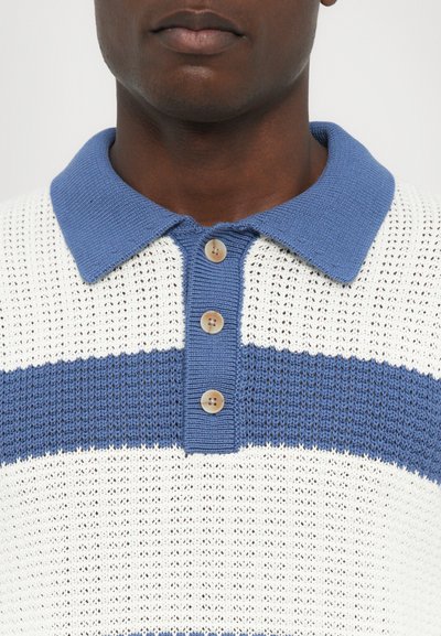 Strikket polo skjorte i hvitt med blå horisontale striper. Har en blå ribbestrikket krage og tre runde knapper ved halsen. Teksturert stoff.