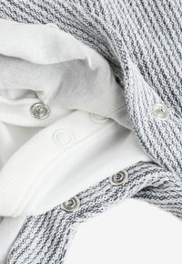 Gros plan sur des vêtements en tricot blanc et gris avec des boutons-pression métalliques partiellement ouverts, révélant des textures de tissus superposés et des attaches.
