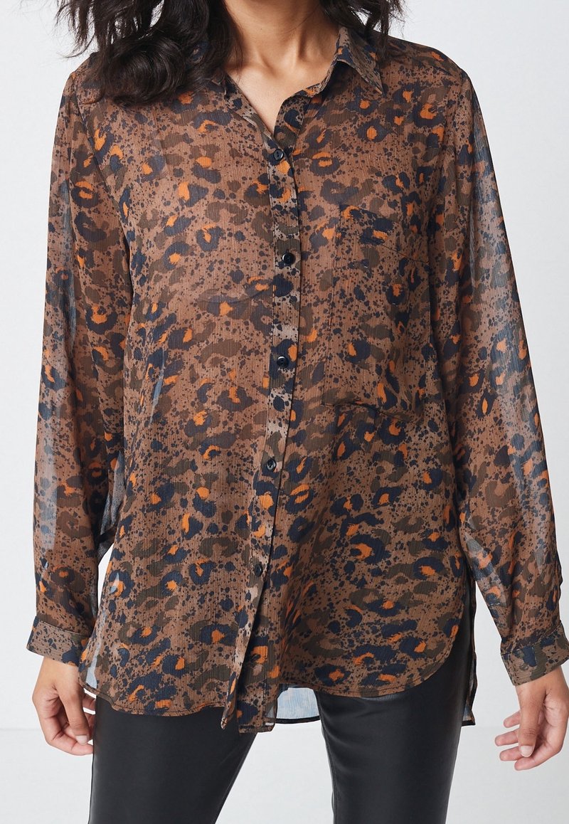 Femme portant une chemise transparente à boutons imprimée léopard marron, orange et noir, manches longues, et un pantalon noir.
