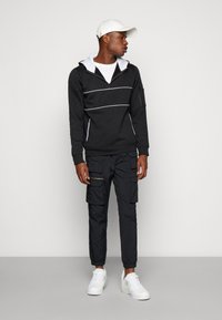 Felpa nera con zip e linee bianche, abbinata a pantaloni cargo neri con tasche con zip. Sneakers bianche completano l'outfit.