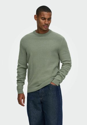 Homme aux cheveux courts porte un pull texturé vert clair et un jean bleu foncé, une main dans la poche, regardant légèrement vers le bas sur le côté.
