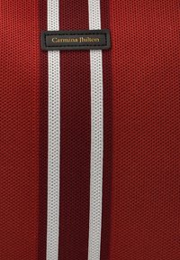 Tela roja con una superficie texturizada, que presenta dos franjas blancas verticales y una etiqueta negra grabada con "Carmina Jhilton" en dorado.