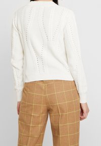 White cable-knit sweater with a textured pattern and a ribbed hem, paired with brown plaid trousers featuring yellow lines.