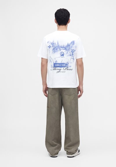 Άνδρας που φοράει λευκό T-shirt με μπλε γραφικό που δείχνει μια καρέκλα σκηνοθέτη και κάμερα, σε συνδυασμό με φαρδιά χακί παντελόνια και αθλητικά παπούτσια.