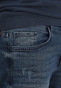 Mørkeblå denim jeans med en baglomme, subtile falmede områder og små slidte detaljer. Stoffet har en glat tekstur.