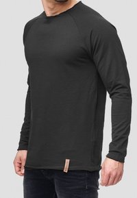 Camisa negra de manga larga con un corte relajado. Presenta un escote deshilachado y un dobladillo lateral, complementada por una pequeña etiqueta de la marca en la parte inferior.