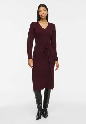 Femme portant une robe bordeaux longue jusqu'aux genoux, à manches longues avec une ceinture nouée et des bottes à talons noires montant aux genoux, se tenant devant un fond uni.