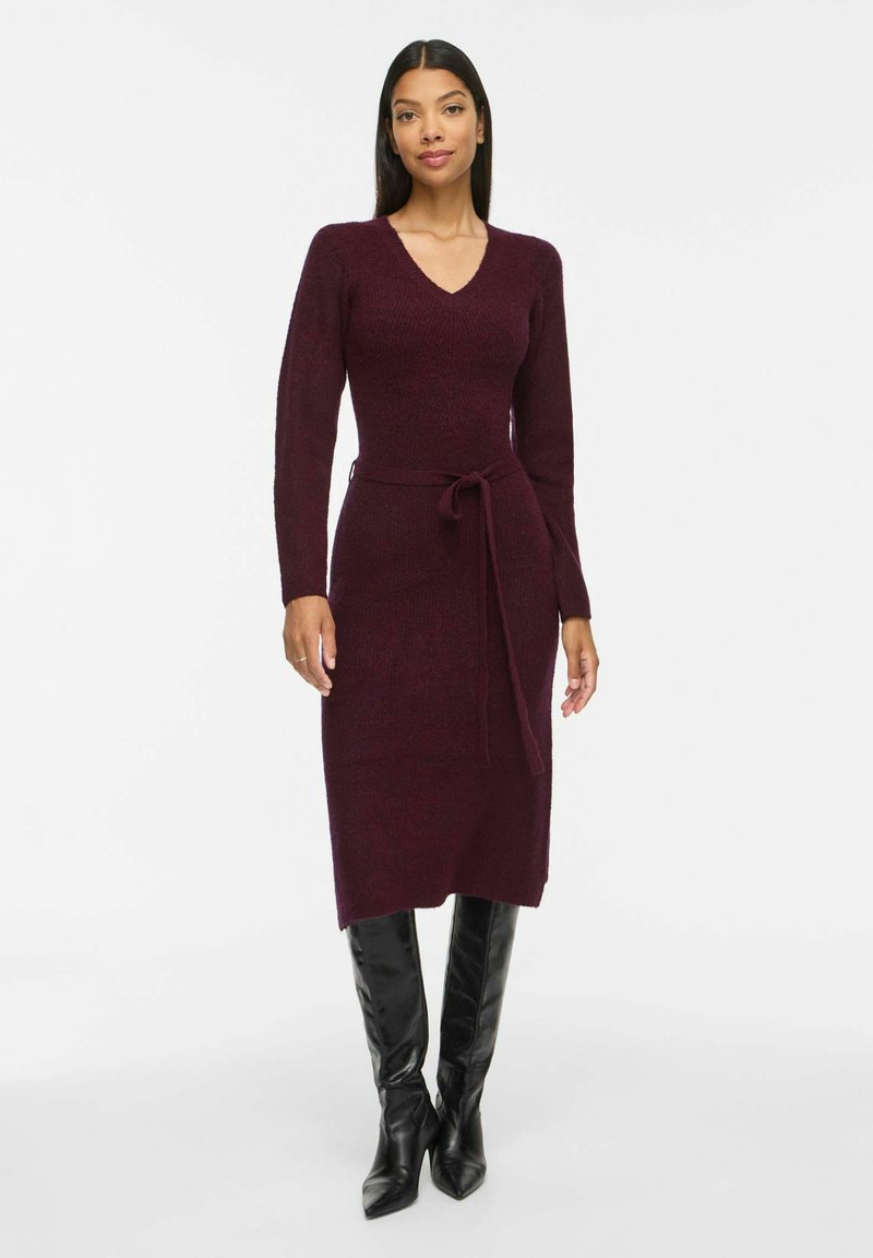 Femme portant une robe bordeaux longue jusqu'aux genoux, à manches longues avec une ceinture nouée et des bottes à talons noires montant aux genoux, se tenant devant un fond uni.